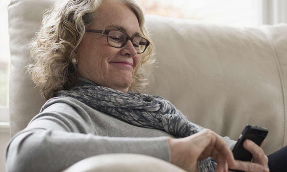 Woman wearing a grey sweater, scarf, and eyeglasses sitting on a couch smiling.